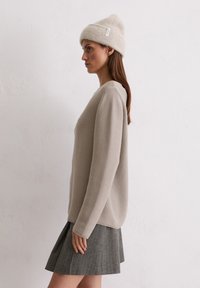 Lichtbeige gebreide trui met een ontspannen pasvorm en geribbelde textuur, gecombineerd met een grijze geplooide rok. Wolachtige beige beanie met een label.