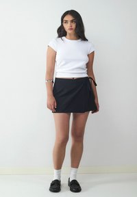 Weißes kurzärmeliges, tailliertes Oberteil kombiniert mit einem schwarzen Wickelrock. Das Outfit hat ein Bindedetail an der Taille und wird mit schwarzen Slippern und weißen Socken kombiniert.