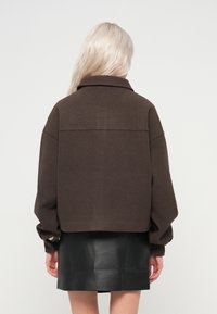 Oseba z dolgimi blond lasmi nosi temno rjav skrajšan plašč in črno usnjeno krilo, pogledana zadaj na enobarvni ozadju.