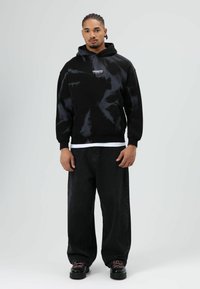 Zwart tie-dye hoodie met een voorzak en geribbelde manchetten, gedragen over een wit shirt. Gecombineerd met losse zwarte jeans en zwarte schoenen.