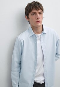 Camicia button-up azzurro chiaro realizzata in tessuto morbido, con colletto classico e vestibilità rilassata, indossata sopra una t-shirt bianca.