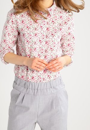 Kobieta w białej, kwiecistej bluzce z długim rękawem z różowymi kwiatami i jasnoszarych spodniach z wysokim stanem, ręce delikatnie splecione na wysokości pasa.