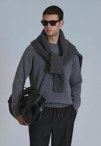 Grijze trui met een geribde sjaal om de hals, gecombineerd met donkere broek. Houdt een zwarte leren tas vast en draagt een zonnebril.