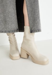 Beige Leder-Ankle-Boots mit einer glatten Textur, quadratischen Zehen und breiten Blockabsätzen, kombiniert mit einem grauen Jeansrock.