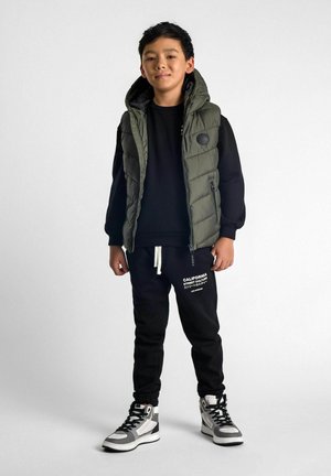 Gilet matelassé vert olive sur un sweat à capuche noir, associé à un pantalon de jogging noir avec du texte blanc, et des baskets montantes avec des accents gris.
