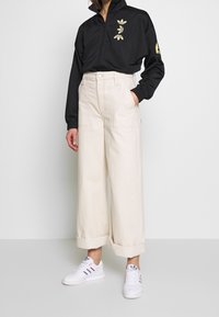 Black zip-up jacket with gold graphic, cream wide-leg pants with rolled cuffs, and white sneakers featuring blue and red stripes.