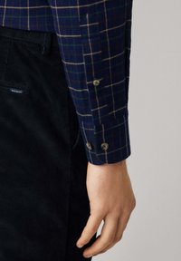 Camicia di flanella a quadri navy con linee verdi e beige, realizzata in tessuto, abbinata a pantaloni in velluto a coste neri. Primo piano della manica della camicia e della mano.