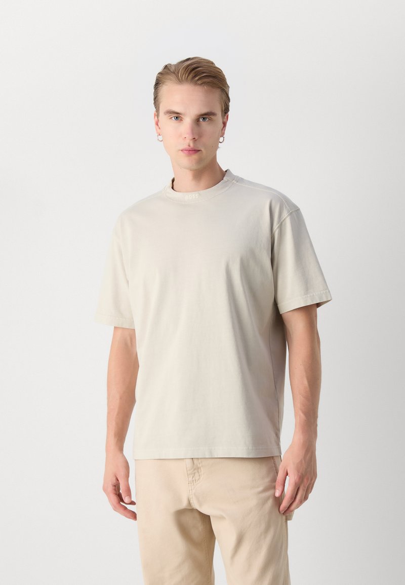 Lichtbeige katoenen t-shirt met korte mouwen, ronde hals en subtiele logodetail bij de kraag. Zachte textuur en een ontspannen pasvorm.