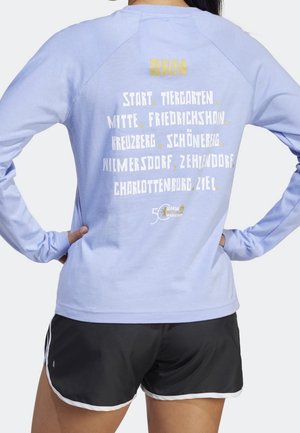 Osoba nosząca jasnofioletową koszulkę z długim rękawem z wymienionymi dzielnicami Berlina i logo maratonu na plecach, połączoną z czarnymi sportowymi szortami.