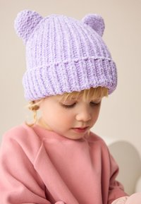 Lilla strikket hue med to afrundede, ørelignende detaljer på toppen. Har tekstureret ribdesignet. Båret af et barn i pink sweatshirt.