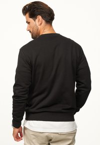 Black crewneck sweatshirt with ribbed texture and cuffs, featuring a relaxed fit and a hem resting at the waist. White shirt visible underneath.