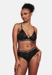 Bralette in pizzo nero con spalline regolabili e banda logo, abbinato a mutandine in pizzo coordinate con una ampia vita nera.