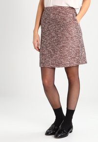 Una gonna corta marrone con texture e un leggero svaso, dotata di tasche laterali e indossata con collant a rete nera e scarpe nere lucide.