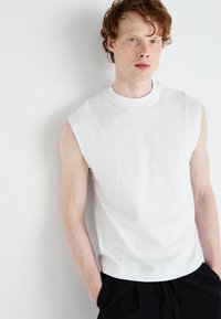 Tricou alb din bumbac, fără mâneci, cu guler rotund și textură netedă, purtat cu pantaloni negri, cu un croi relaxat și un design minim.