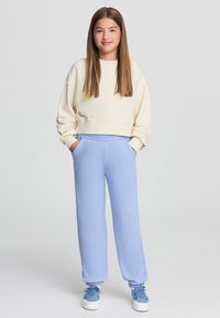 Felpa corta beige chiaro abbinata a pantaloni della tuta blu chiaro. L'outfit presenta polsini elasticizzati e tasche, su uno sfondo semplice.