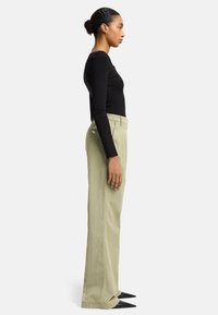 Zwarte aansluitende top met lange mouwen gecombineerd met lichtgroene wijde broek; model draagt zwarte puntige schoenen; neutrale achtergrond.