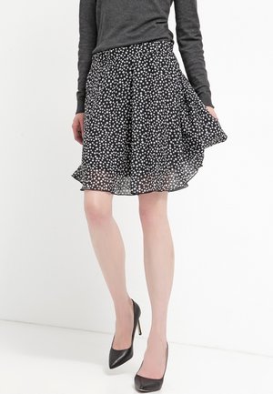 Vrouw draagt een zwart-witte polka dot rok en een donkergrijze top met lange mouwen, houdt de zoom van de rok vast, gecombineerd met zwarte pumps.