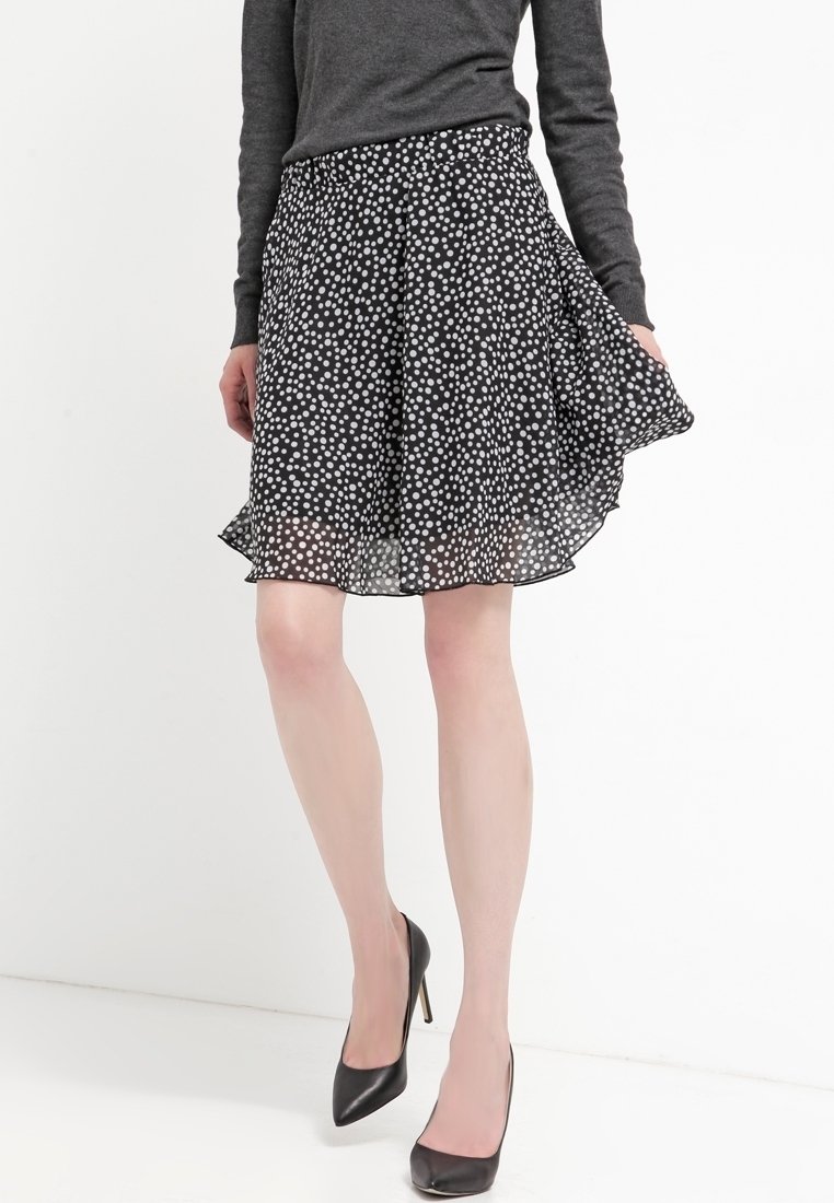 Femme portant une jupe à pois noirs et blancs et un haut à manches longues gris foncé, tenant l'ourlet de la jupe, assortie à des talons noirs.