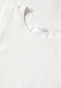 Witte katoenen tanktop met een ronde halslijn. Heeft een subtiele textuur en een label met de naam "STREET ONE STUDIO."