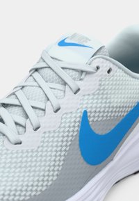 Šedá sportovní bota s mesh svrškem, modrým logem Nike, bílými akcenty a texturovanými tkaničkami pro lepší grip. Obsahuje polstrovanou podrážku pro pohodlí.