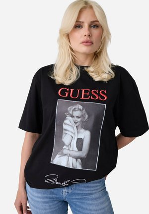 Femme blonde porte un t-shirt noir Guess avec un lettrage rouge et une photo en noir et blanc de Marilyn Monroe tenant un châle en fourrure, associé à un jean bleu.