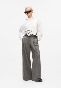 White hoodie with black "Karl" lettering, paired with gray wide-leg trousers in a textured fabric, and large black angular sunglasses.