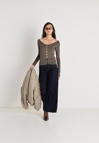 Γυναίκα με ελαιοπράσινη μπλούζα με κουμπιά, σκούρες παντελόνες φαρδιάς γραμμής, γυαλιά ηλίου, κρατώντας μπεζ μπουφάν, περπατώντας μπροστά σε λευκό φόντο.