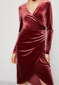 Robe portefeuille en velours d'un rouge profond avec des manches longues, taille froncée et ourlet asymétrique, sur une personne debout montrée de la nuque aux genoux.