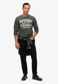 Grijze lange mouw shirt met "Superdry" tekst, zwarte jeans, geruite shirt om de taille geknoopt en zwarte veterschoenen.