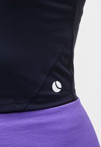 Μαύρο εφαρμοστό τοπ με μικρό λευκό λογότυπο τεννίστας κοντά στη ραφή του πλάι, σε συνδυασμό με υφασμάτινο μοβ κάτω ένδυμα.