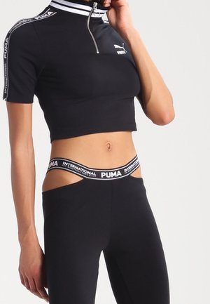 Kobieta w czarnej krótkiej bluzie Puma zapinanej na zamek i dopasowanych legginsach z wysokim, markowym paskiem odsłaniającym brzuch.