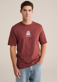 Maronfarvet bomulds T-shirt med korte ærmer og rund halsudskæring, der har en hvid grafik af en bygning med teksten "Hotel Di Sonos 1998" trykt nedenfor.