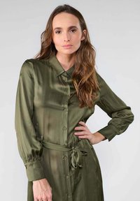 Robe verte olive à manches longues en texture soyeuse, avec une taille cintrée et un col. Présente un léger éclat et des poignets froncés.