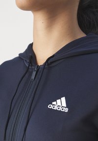 Navy zipepová mikina vyrobená z měkkého materiálu, s černým zipem, bílým logem Adidas a nastavitelnou kapucí se šňůrkami.