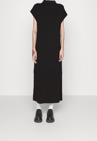 Schwarzes, geripptes Maxikleid mit hohem Kragen und kurzen Ärmeln, mit seitlichen Schlitzen, getragen mit schwarzen klobigen Schuhen und weißen Socken.