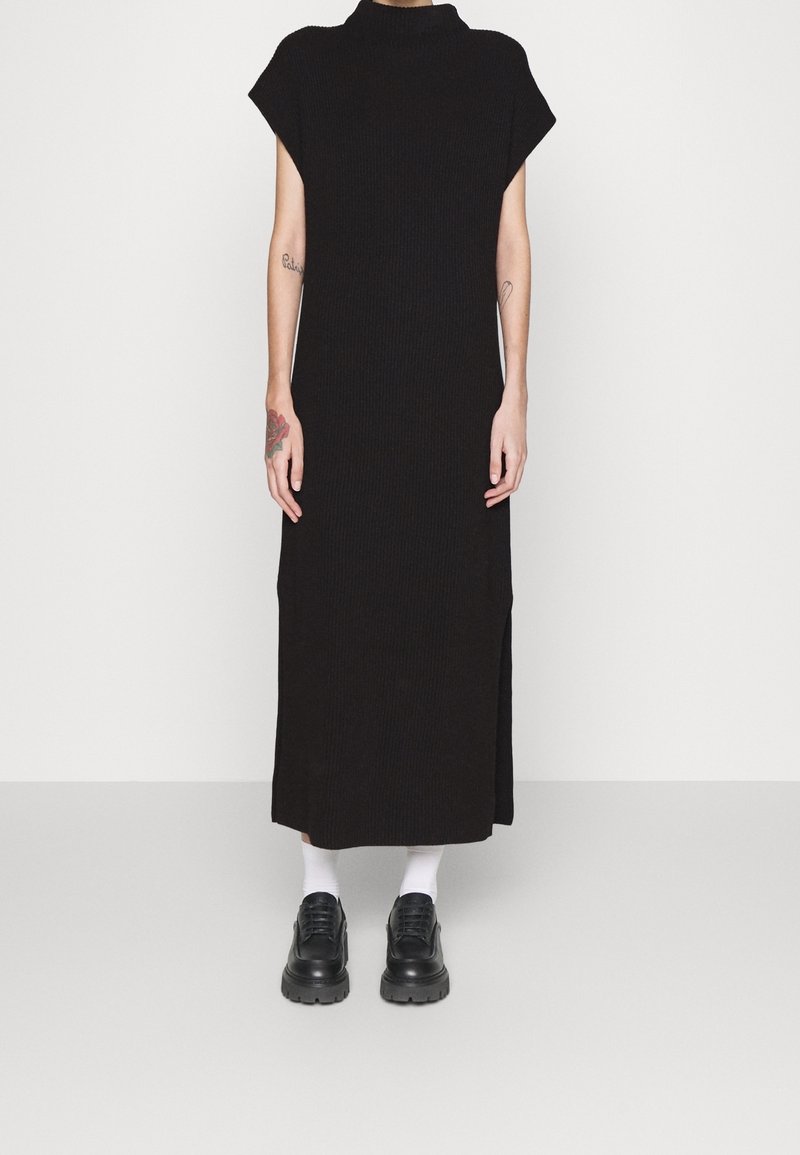 Schwarzes, geripptes Maxikleid mit hohem Kragen und kurzen Ärmeln, mit seitlichen Schlitzen, getragen mit schwarzen klobigen Schuhen und weißen Socken.