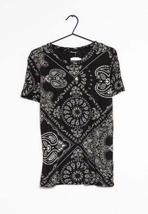 Musta lyhythihainen t-paita, jossa valkoisia paisley- ja geometrisia kuvioita, pyöreä pääntie ja pehmeä kangas, roikkuu mustassa naulassa.