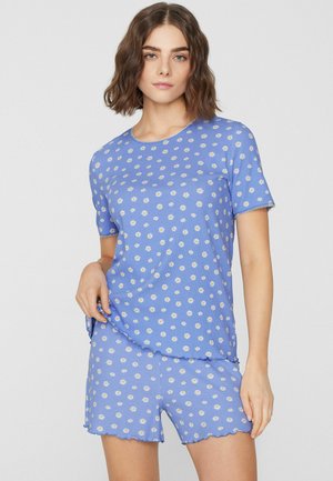 Femme portant un haut à manches courtes bleu clair et un short assorti avec des motifs de marguerites blanches, posant devant un fond clair uni.