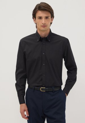 Junger Mann trägt ein eng anliegendes schwarzes langärmeliges Hemd mit Knöpfen und marineblaue Hose, steht vor einem schlichten hellen Hintergrund.