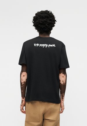 Άτομο με σκούρα σγουρά μαλλιά, που φοράει μαύρο μπλουζάκι με λευκή επιγραφή «THE NORTH FACE» στην πλάτη και καφέ παντελόνι, στέκεται μπροστά σε ένα απλό φόντο.