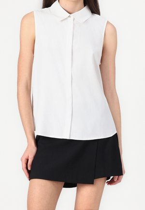 Blouse blanche sans manches boutonnée avec col, portée avec une mini-jupe asymétrique noire sur une personne debout.