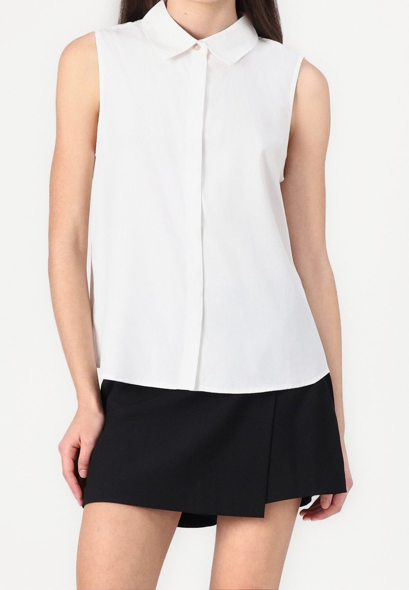 Blouse blanche sans manches boutonnée avec col, portée avec une mini-jupe asymétrique noire sur une personne debout.