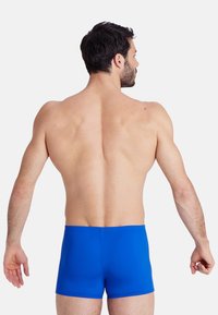 Bañador azul hecho de una tela elástica, con un diseño simple y elegante. La vista trasera muestra una forma ajustada y una textura suave.