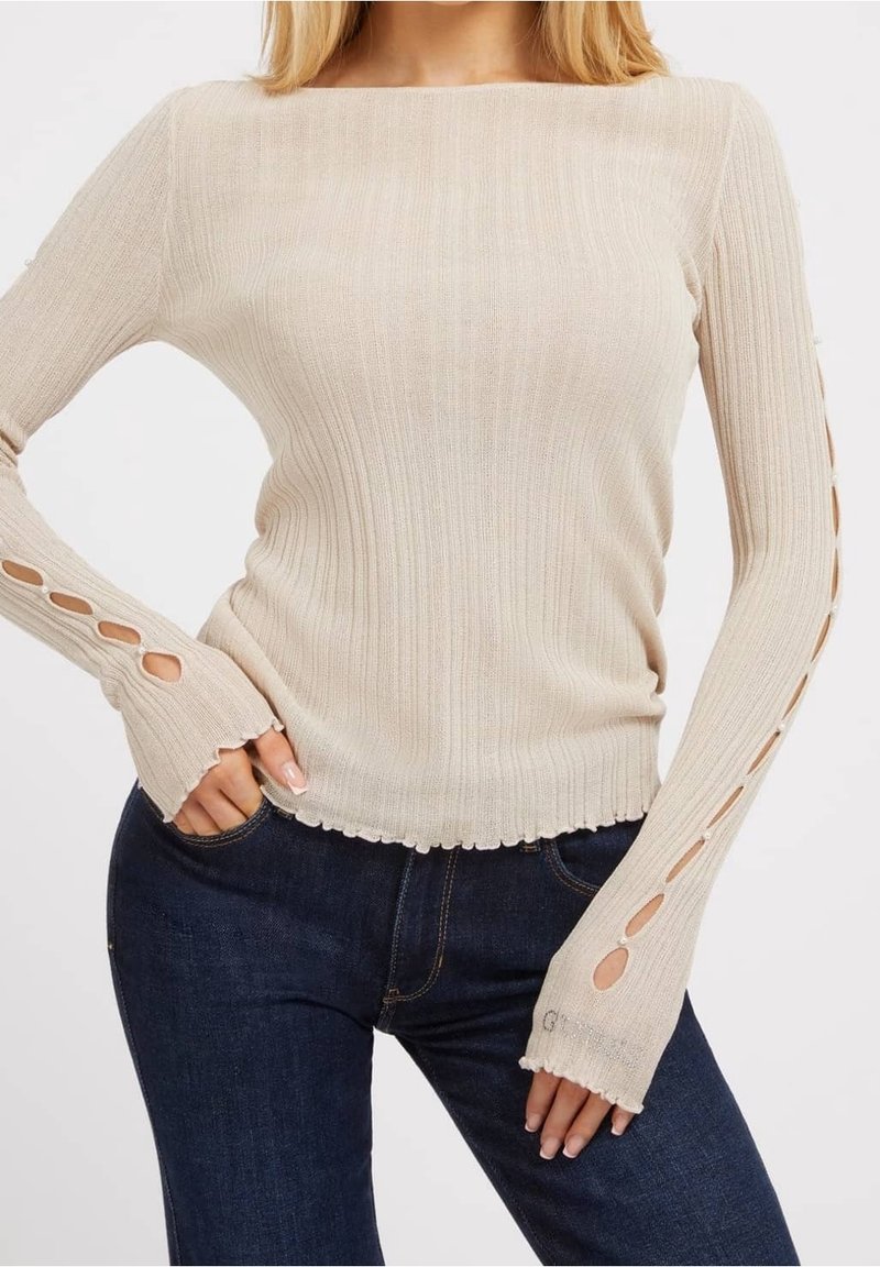 Top beige a maniche lunghe a costine con orlo smerlato e dettaglio aperto sulle maniche. Il tessuto ha una texture morbida, che si abbina a jeans blu scuro.
