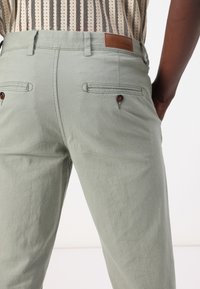 Pantalons vert clair avec un tissu texturé, dotés de deux poches arrière avec des boutons marron et d'une pièce en cuir marron à la taille.