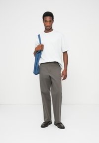 Γκρι παντελόνια, λευκό φαρδύ μπλουζάκι και σκούρα παπούτσια. Μια μπλε τσάντα ώμου κρέμεται στον ώμο. Απλό φόντο.