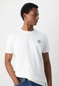 Camiseta blanca de algodón con cuello redondo y mangas cortas, con un emblema de león texturizado en el pecho y un corte relajado.