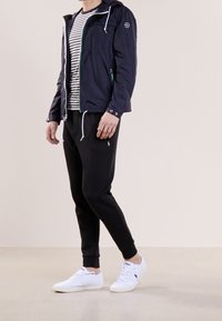 Marinblå vindtät jacka med huva, vita dragsnören, sidofickor, bärs över en randig skjorta och kombineras med svarta joggers och vita sneakers.