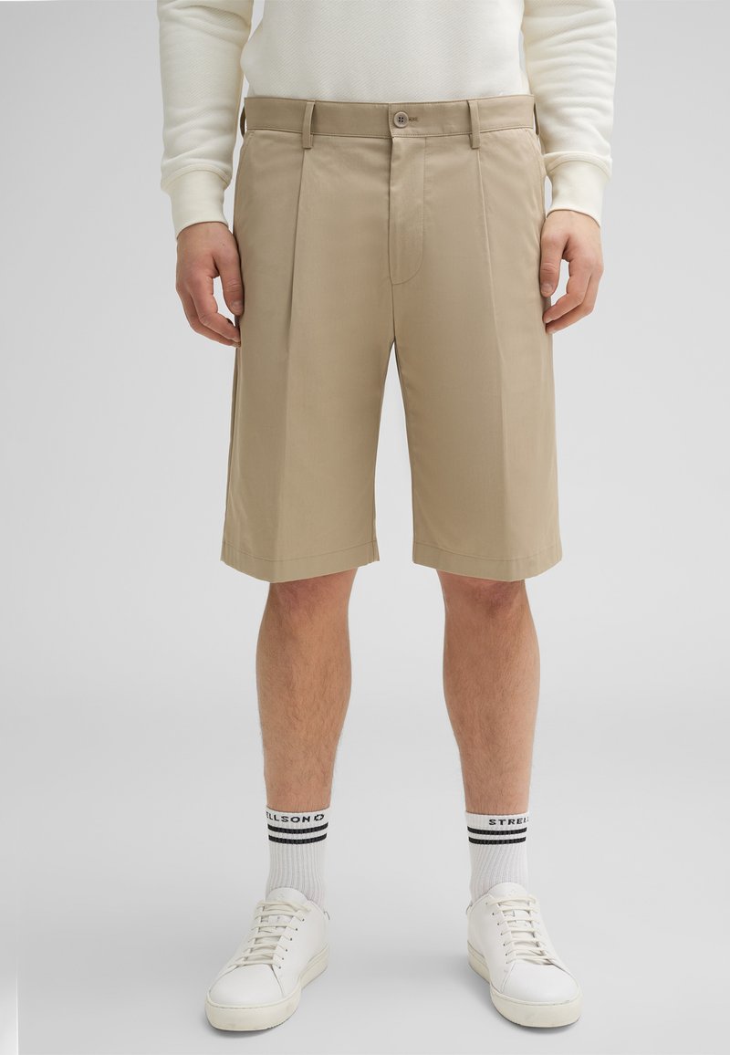Pantaloni beige su misura con pieghe frontali, tessuto misto cotone, taglio fino al ginocchio, abbinati a sneakers bianche e calzini a righe.