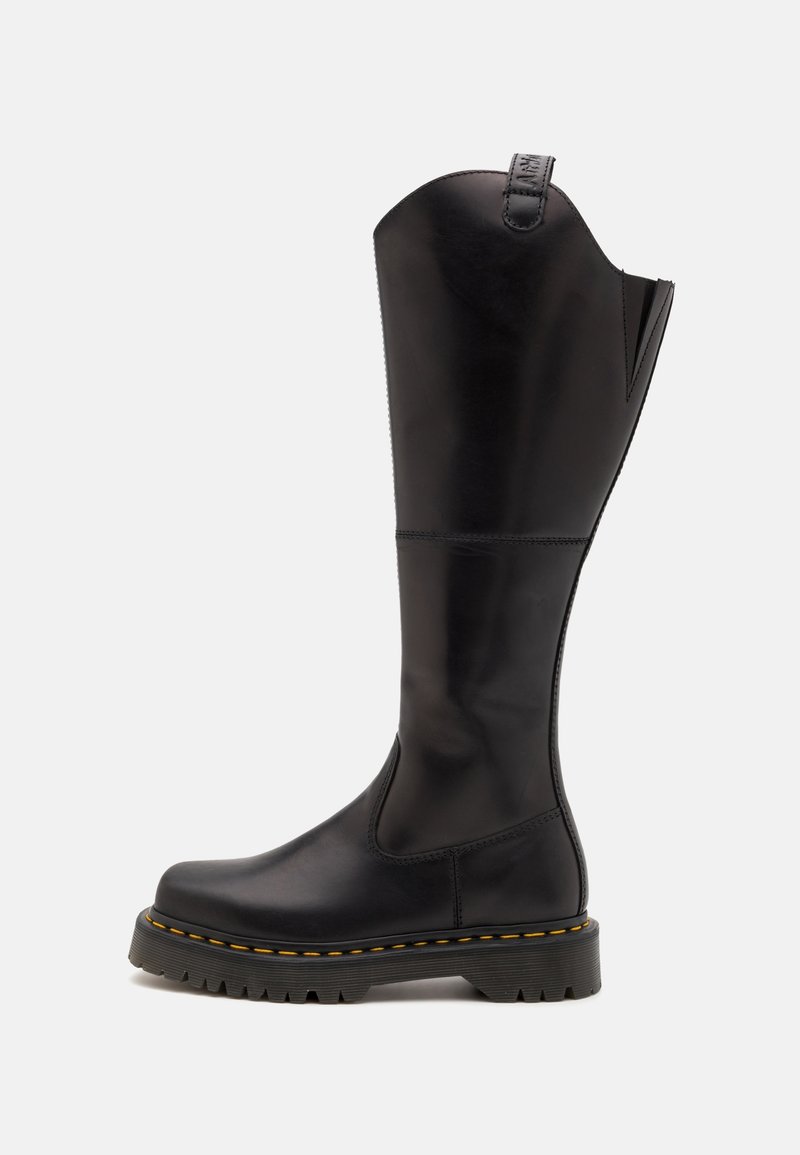 Botte noire en cuir montant au genou avec semelle en caoutchouc épaisse, couture jaune et languette de tirage en haut sur un fond uni.